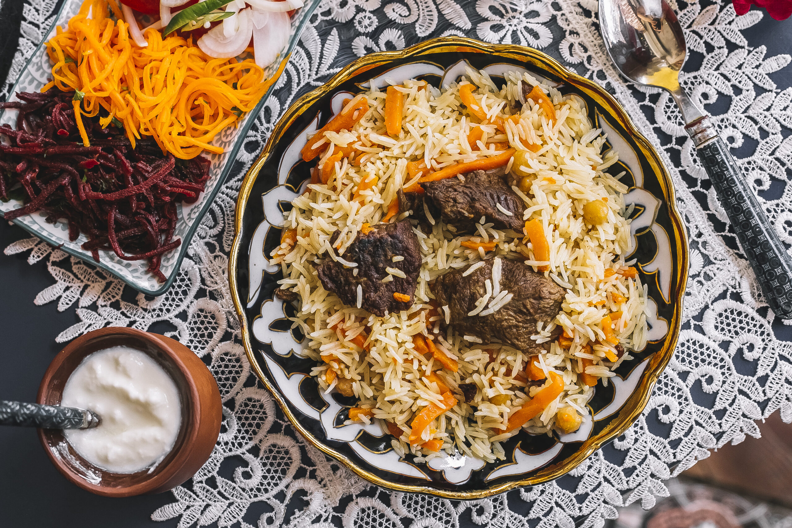 "Top view of a plate of rice with carrots and pieces of lamb, served on a decorative tablecloth alongside a bowl of yogurt and a dish of sliced vegetables."